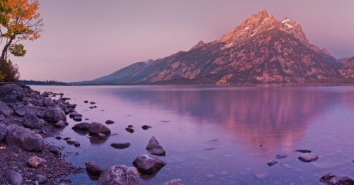 A serene lake reflects a mountain at dusk, surrounded by rocky shores and autumn foliage.