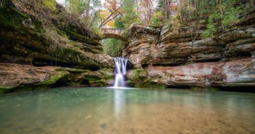 A serene waterfall cascades into a clear pool, surrounded by lush greenery and rocky cliffs, with a bridge in the background.
