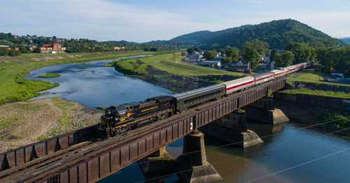 A vintage train crosses a bridge over a river, surrounded by green hills and a small town in the background.