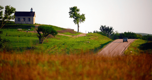 A scenic view of a rural road winding past a small stone church on a grassy hill, with trees in the background.