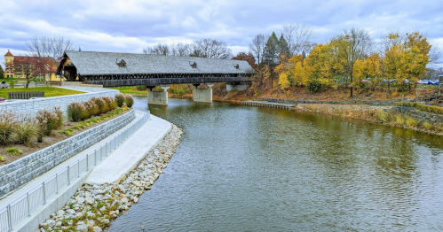 A covered bridge spans a river, surrounded by autumn trees and a landscaped pathway along the water's edge.