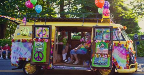 A colorful, decorated yellow van with balloons, flowers, and peace signs, featuring two people inside, enjoying a festive atmosphere.