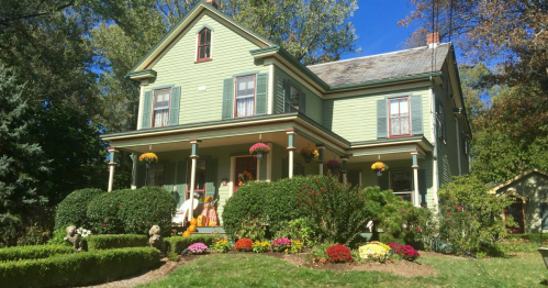 A charming green Victorian house with a porch, surrounded by colorful flowers and well-maintained shrubs.
