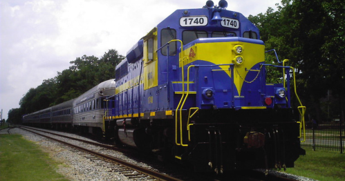 A blue and yellow train engine with passenger cars parked on a railway track under a cloudy sky.