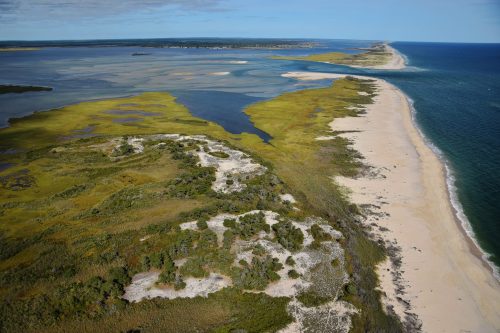 Aerial view of a coastal landscape featuring sandy beaches, green marshlands, and winding waterways under a clear blue sky.