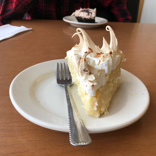 A slice of creamy cake topped with whipped cream and decorative swirls, served on a white plate with a fork.