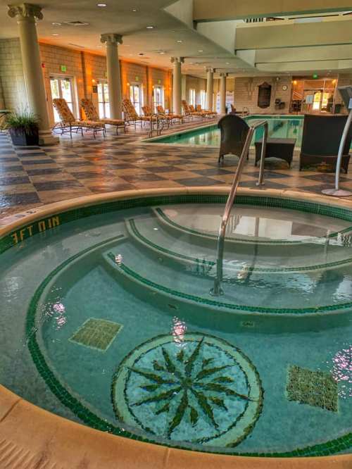 A serene indoor pool area featuring a hot tub with a decorative tile design and lounge chairs in the background.