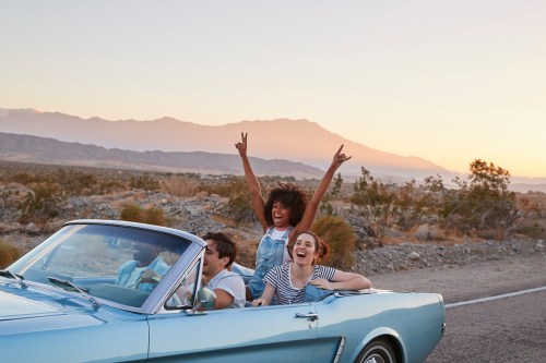 Friends cheer in vintage drop-top car on golden-hour highway