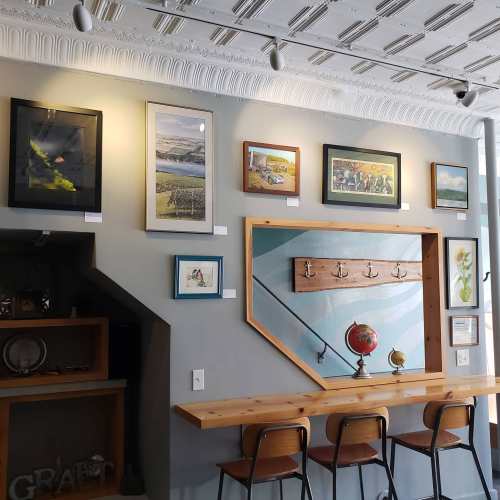 A cozy café wall featuring various framed artworks and a wooden bar with stools, illuminated by natural light.