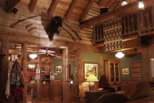 Cozy rustic interior of a lodge with wooden beams, a bison head mount, and warm lighting.