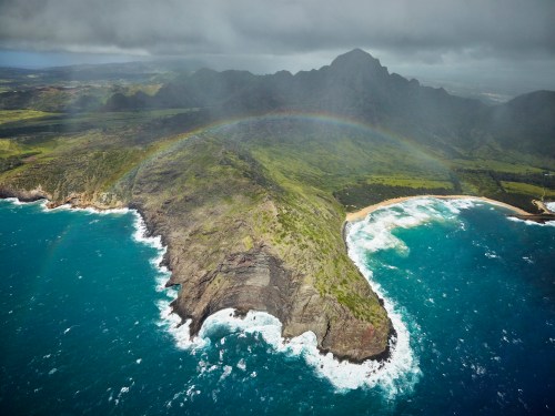 Aerial view of a lush green coastline with a rainbow arching over a rocky peninsula and crashing waves below.