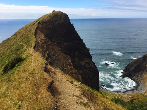 A rugged coastal cliff with grassy slopes overlooking the ocean and waves crashing against the rocks below.