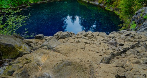 A rocky ledge overlooking a deep blue pool surrounded by lush greenery and trees reflecting in the water.