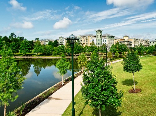 A serene park scene featuring a calm lake, green trees, and modern buildings under a blue sky with fluffy clouds.