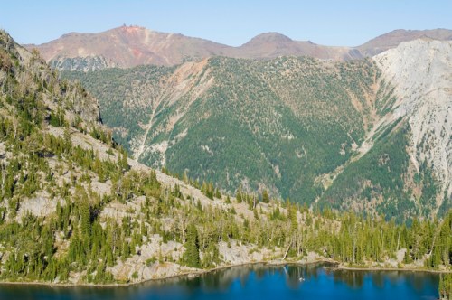 A serene mountain landscape featuring lush green forests, rocky peaks, and a calm blue lake in the foreground.