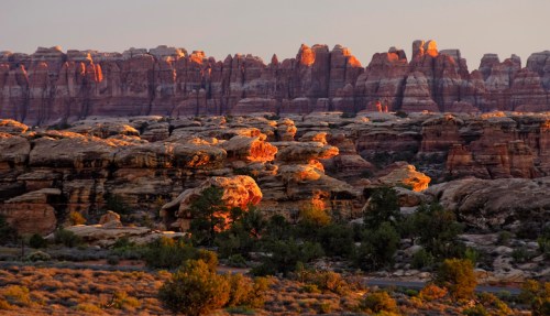 Sunset over rugged rock formations and cliffs, casting warm light on the landscape in a desert setting.