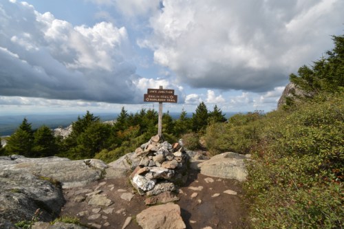 A mountain trail sign stands among rocks and trees, with a cloudy sky in the background.