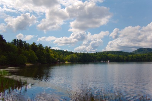 A serene lake surrounded by lush greenery and mountains, under a bright blue sky with fluffy white clouds.
