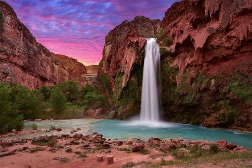 A stunning waterfall cascades into a turquoise pool, surrounded by red rock cliffs and a colorful sunset sky.