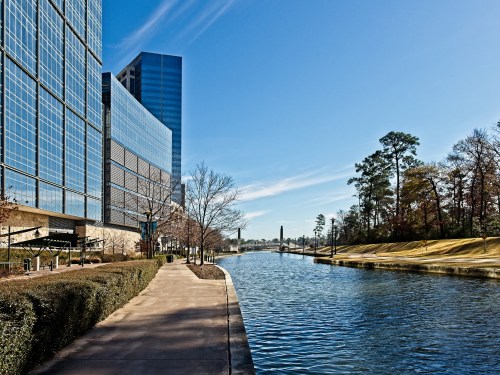 A serene waterway lined with trees and modern buildings under a clear blue sky.