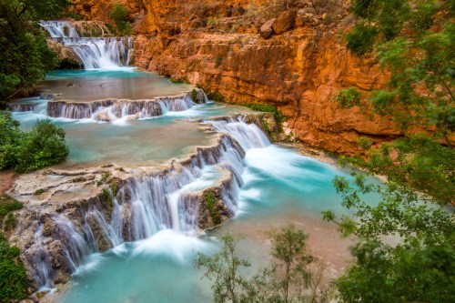 A serene landscape featuring cascading waterfalls over rocky tiers, surrounded by lush greenery and red rock formations.