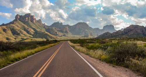 A winding road leads through lush greenery, with dramatic mountains and a cloudy sky in the background.