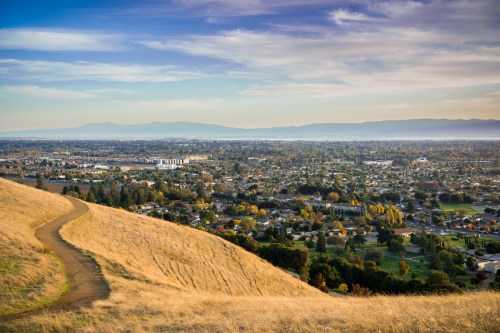 A scenic view of a valley with a winding path, surrounded by hills and a cityscape under a clear blue sky.