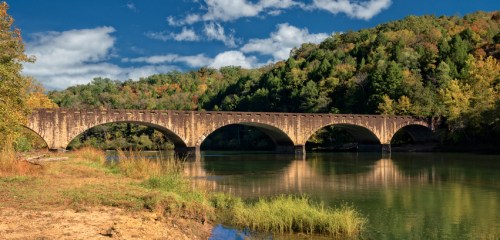 A stone bridge arches over a calm river, surrounded by lush greenery and a blue sky with scattered clouds.