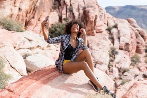 A woman with curly hair sits on a rock, smiling and taking a selfie in a scenic outdoor landscape.