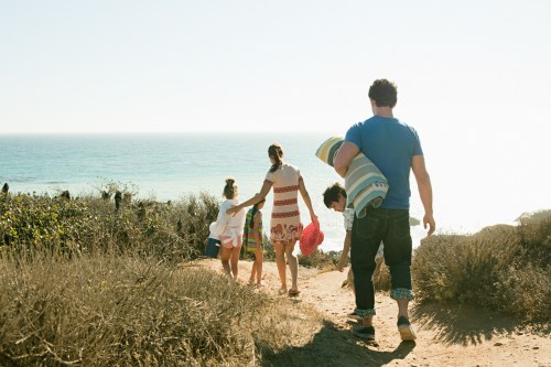 A family walks along a coastal path, enjoying a sunny day by the ocean, with one child being carried on an adult's back.