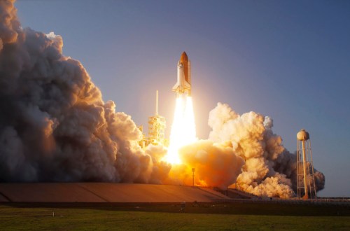 A rocket launches into the sky, surrounded by clouds of smoke and flames against a clear blue backdrop.