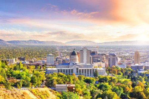 A panoramic view of a city skyline at sunset, featuring buildings, trees, and mountains in the background.