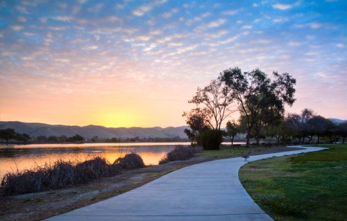 A serene lakeside path at sunrise, with trees and mountains in the background under a colorful sky.
