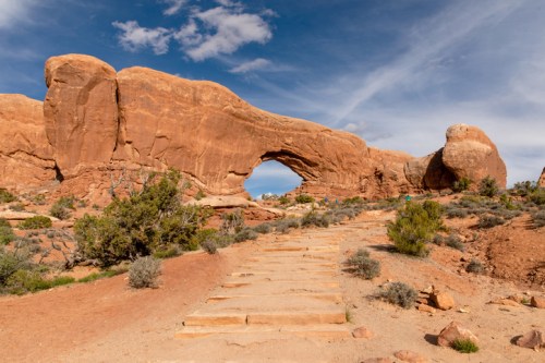 A natural rock arch stands tall against a blue sky, surrounded by desert vegetation and a stone pathway leading up to it.