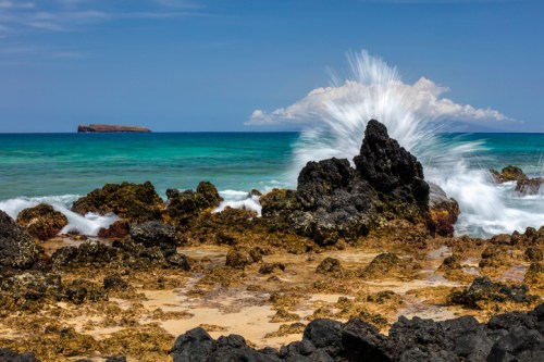 Waves crashing against rocky shore with clear blue water and distant island under a bright sky.