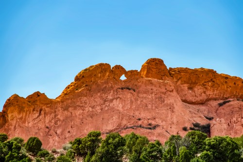 A rocky cliff with a natural arch, surrounded by green shrubs under a clear blue sky.