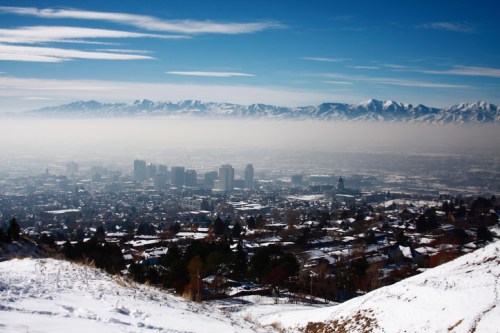 A snowy landscape overlooks a city shrouded in fog, with mountains in the background under a clear blue sky.
