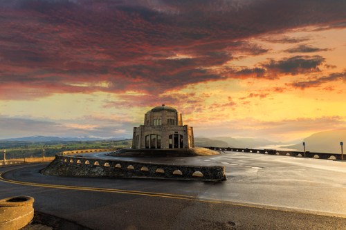 A historic monument on a winding road, set against a vibrant sunset sky with clouds and distant hills.