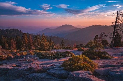 A serene mountain landscape at sunset, featuring rocky terrain, trees, and a colorful sky with clouds.