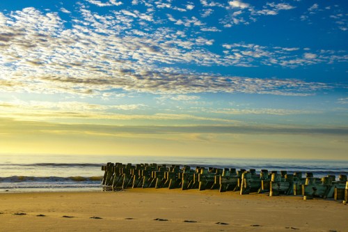 A serene beach scene at sunrise, featuring wooden posts along the shore and a sky filled with clouds.
