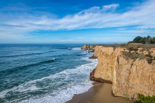 A scenic view of rugged cliffs meeting the ocean under a blue sky with scattered clouds. Waves crash onto the shore.