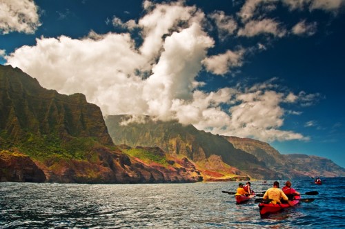 Kayakers paddle along a scenic coastline with dramatic cliffs and vibrant clouds under a blue sky.