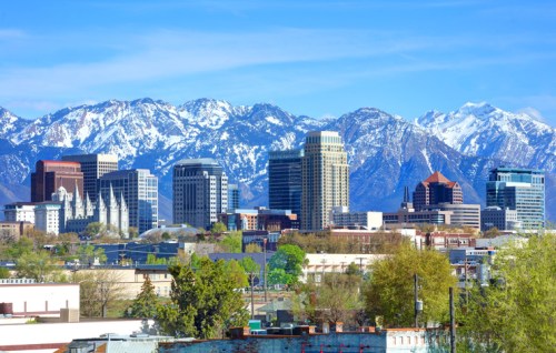 A city skyline with modern buildings in front of snow-capped mountains under a clear blue sky.