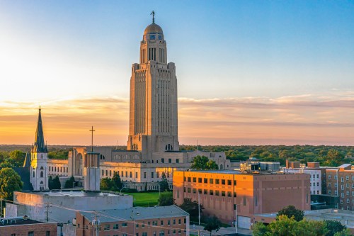 A skyline view featuring a tall building with a clock tower at sunset, surrounded by trees and smaller structures.