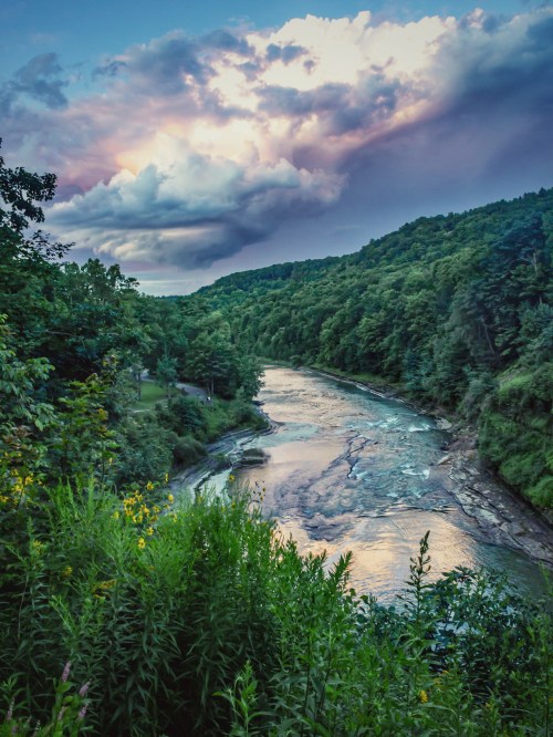 A serene river winds through lush green hills under a dramatic sky with clouds at sunset.