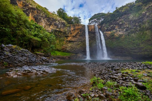 A scenic waterfall cascades into a serene pool, surrounded by lush greenery and rocky terrain under a cloudy sky.
