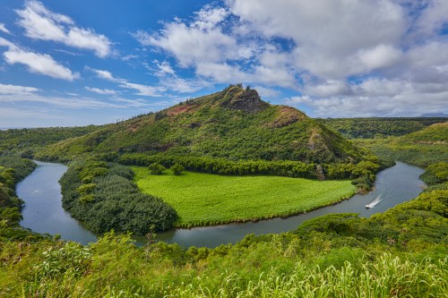 Lush green hills surround a winding river under a blue sky with scattered clouds, creating a serene landscape.