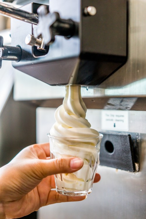 A hand holds a clear cup as soft serve ice cream is dispensed from a machine.