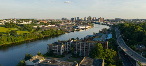 Aerial view of a river with a city skyline in the background, surrounded by greenery and industrial buildings.