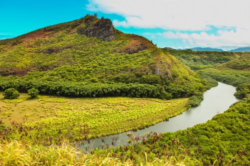 Lush green hills surround a winding river under a bright blue sky, creating a serene natural landscape.
